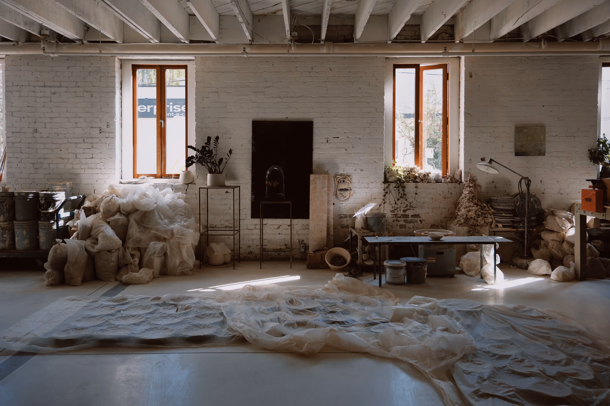 Art studio with white brick walls and two windows letting in natural light, scattered with bags of raw materials covered with translucent plastic sheets, metal tables holding various containers, plants on stands, a floor lamp, and pottery items.