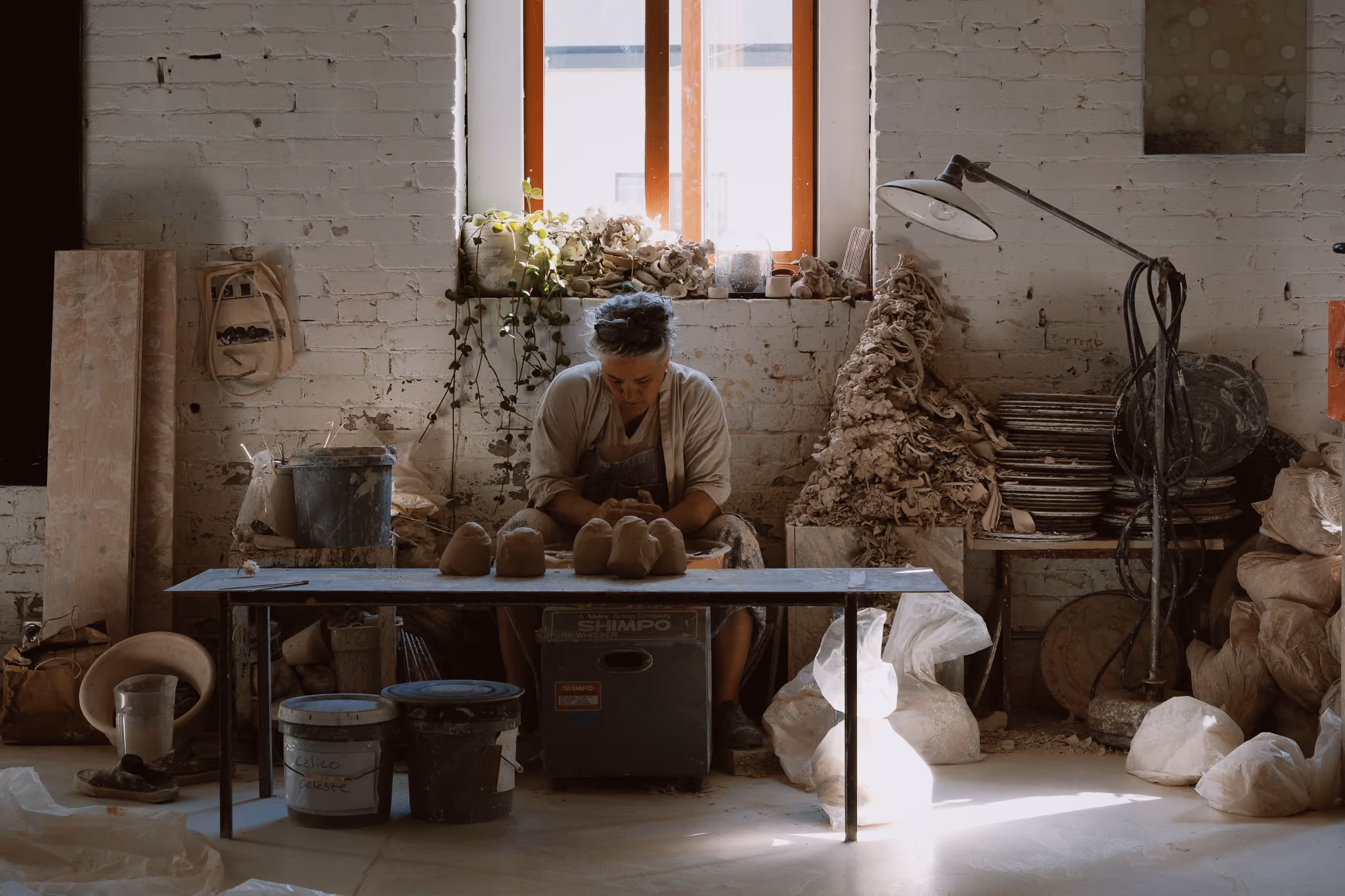 A person working with clay in a pottery studio, seated at a table with several small clay sculptures in front of them, surrounded by pottery tools, containers, and piles of clay scraps, with sunlight streaming through a window behind.