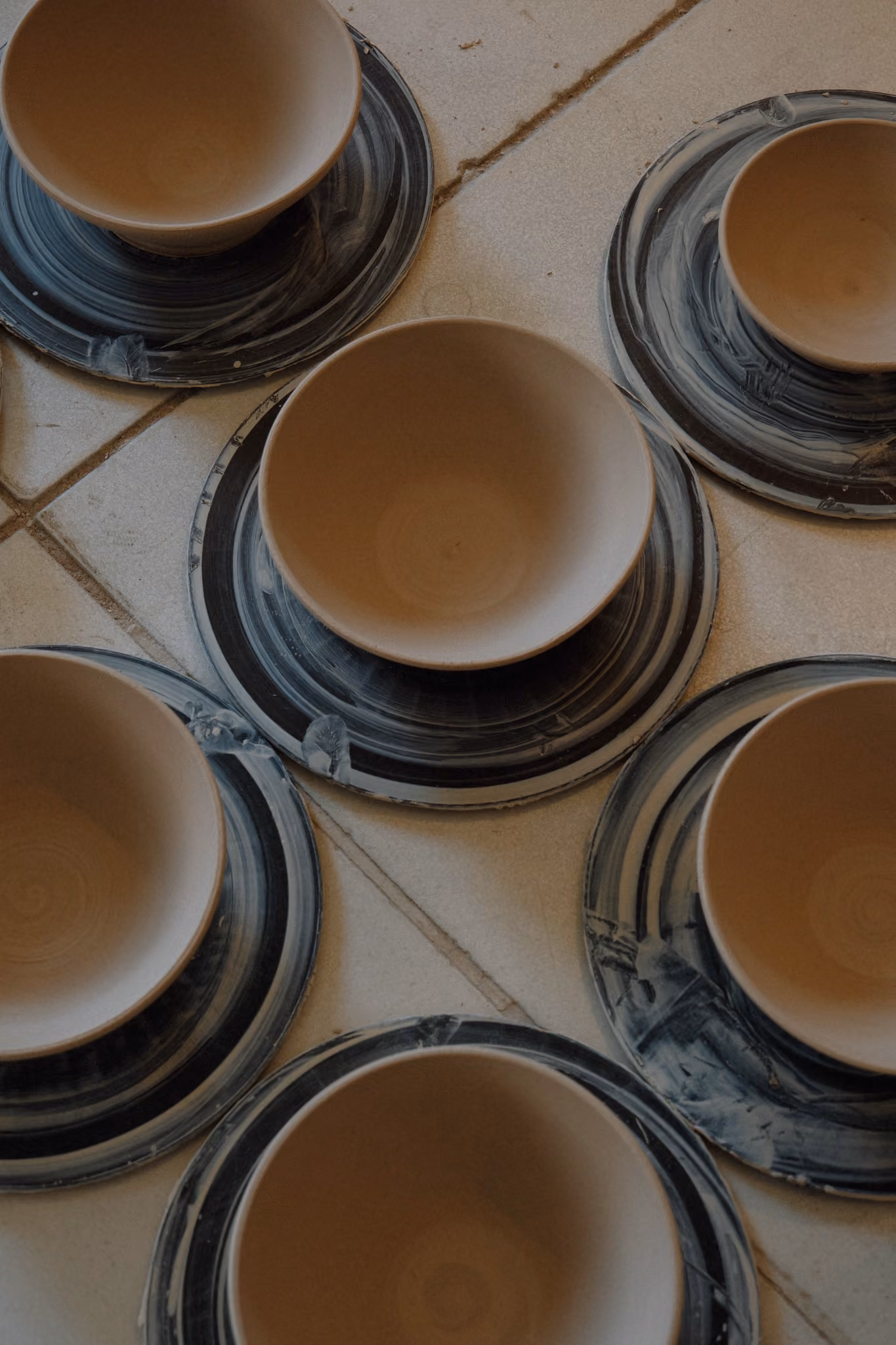 Several unglazed ceramic bowls placed upside down on circular pottery wheels with dark and light swirl patterns, arranged on a tiled floor.
