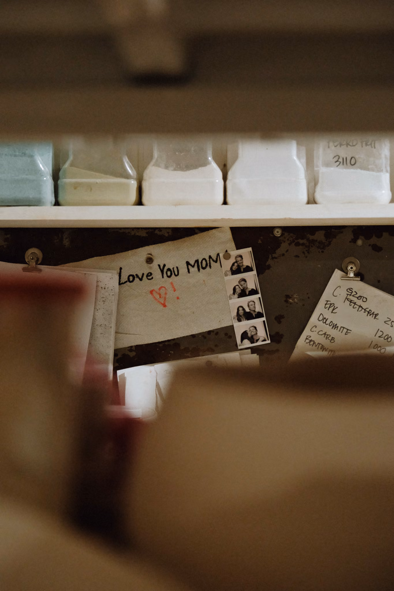 A bulletin board with a handwritten note saying 'Love You MOM' with a red heart and exclamation mark, accompanied by a photo booth strip of a couple making faces, and papers with handwritten notes pinned around it. Above the bulletin board are several translucent containers filled with powders.
