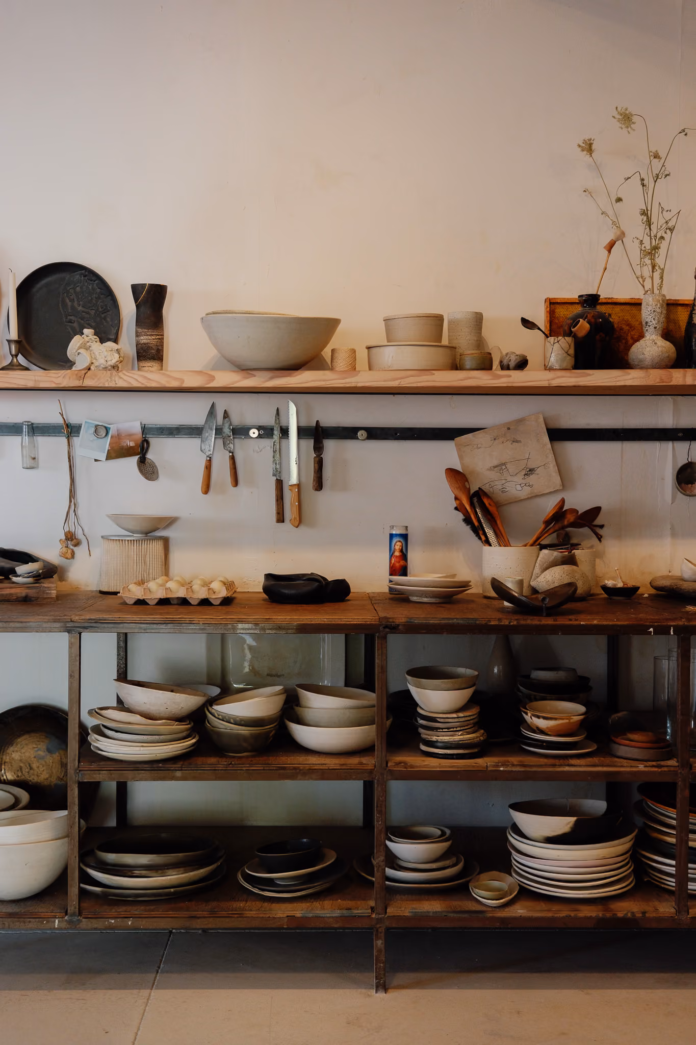 Rustic wooden shelves filled with various ceramic bowls, plates, and pottery pieces, a row of knives hanging on a metal strip above, wooden spoons in containers, and decorative dried plants in vases.