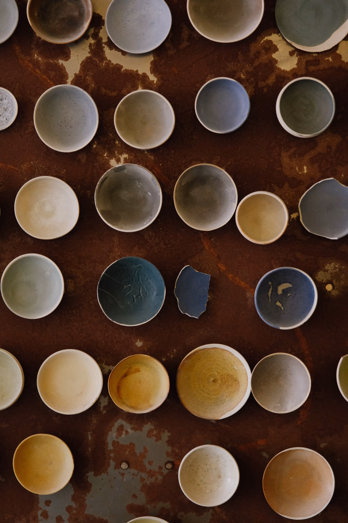 Overhead view of various empty ceramic bowls in neutral tones, including beige, gray, and blue, some with slight texture, arranged on a rusty brown metal surface.