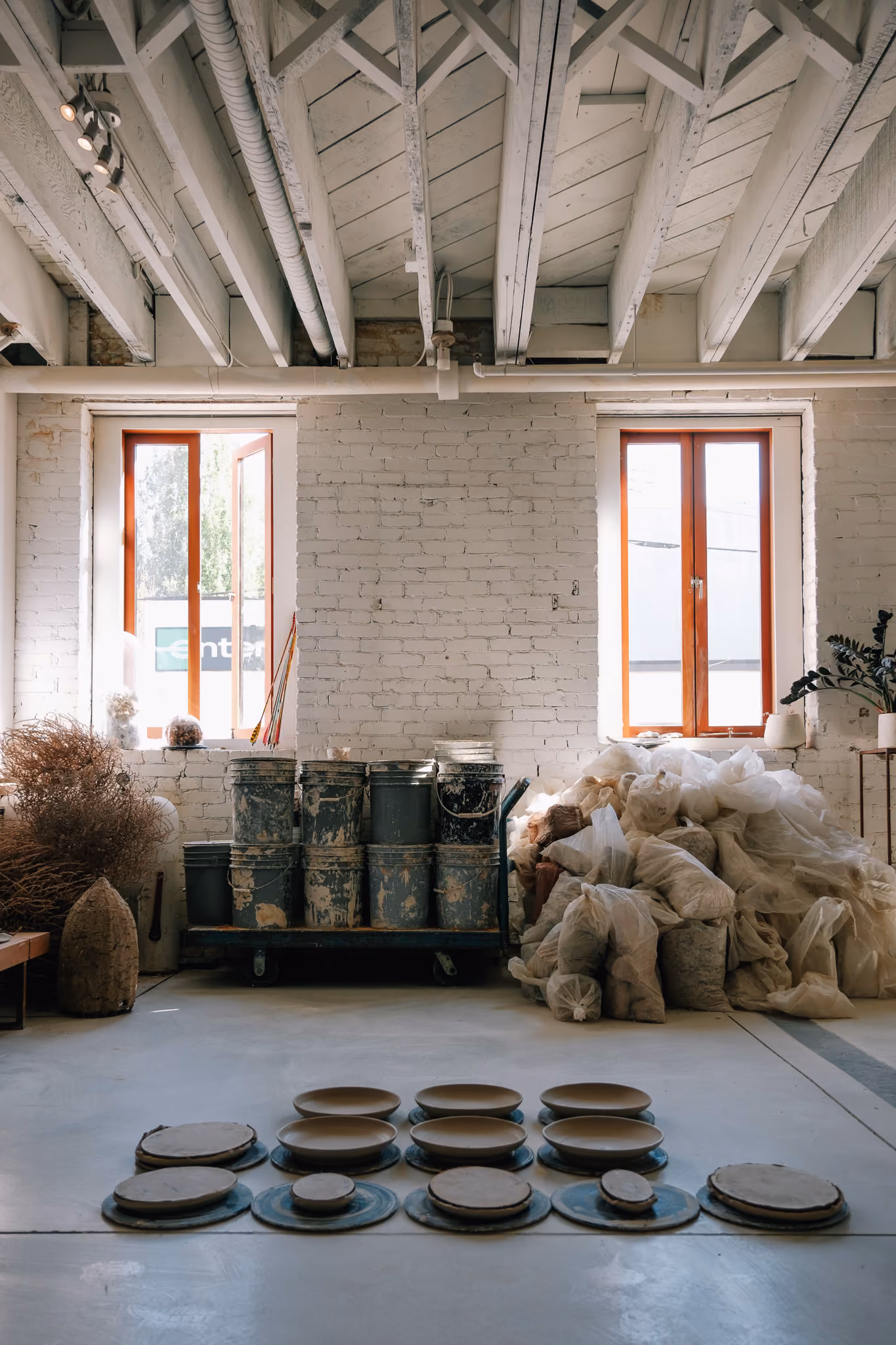 Interior of an art studio with white painted brick walls and exposed ceiling beams, featuring two windows with reddish-brown frames. Below the windows are stacked weathered buckets and a large pile of white bags. In the foreground, unglazed pottery pieces are arranged on circular metal bases on the floor.