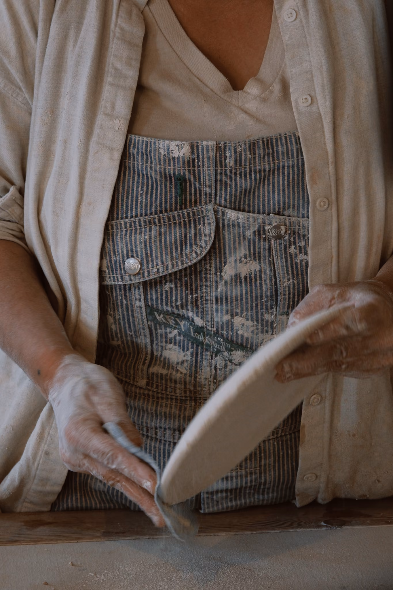 Person wearing a striped denim apron and a beige shirt dusting a round, flat dough with flour, hands covered in flour, working on a wooden surface.