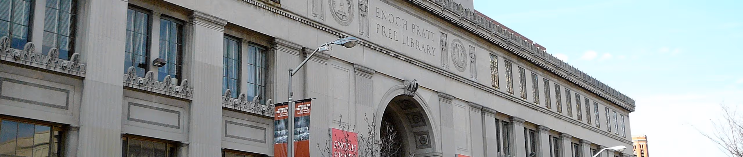 Exterior of a large, historical building, identified as the Enoch Pratt Free Library. The architectural style features classical elements, including decorative details on the facade and tall windows. Banners are visible hanging from the building, and the sky is clear, with a hint of urban surroundings in the background. The structure suggests a prominent and well-established institution.