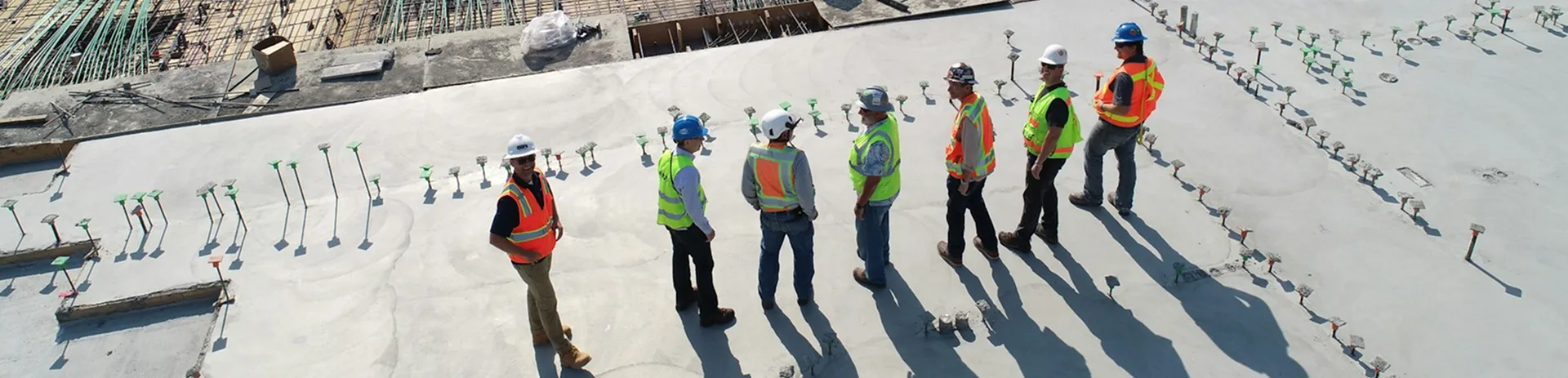 Seven construction workers wearing safety vests and helmets stand on a concrete slab at a construction site, discussing the project as the sun casts long shadows.