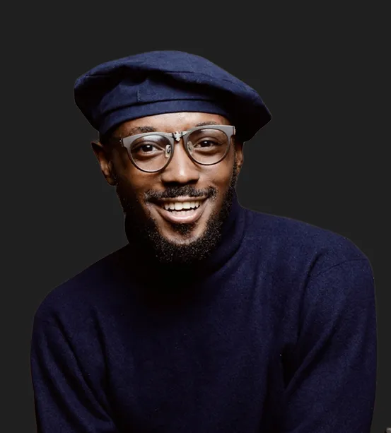 Smiling man wearing a navy blue beret, glasses, and a navy blue turtleneck sweater against a dark background.