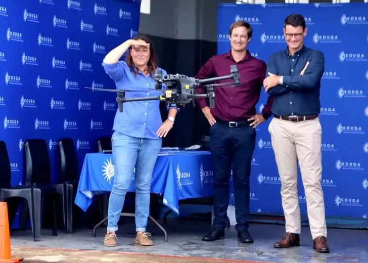 Three people standing in front of blue banners with 'Kouga' logos, with a drone flying in front of them; one woman shielding her eyes while two men smile.