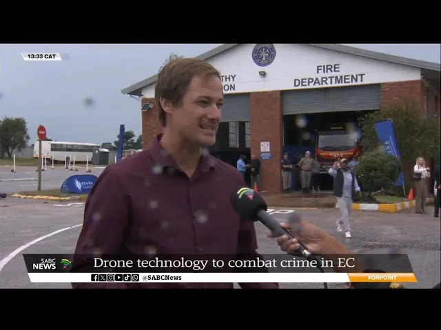 Man in maroon shirt being interviewed outdoors in front of a fire department building with people and a fire truck in the background.