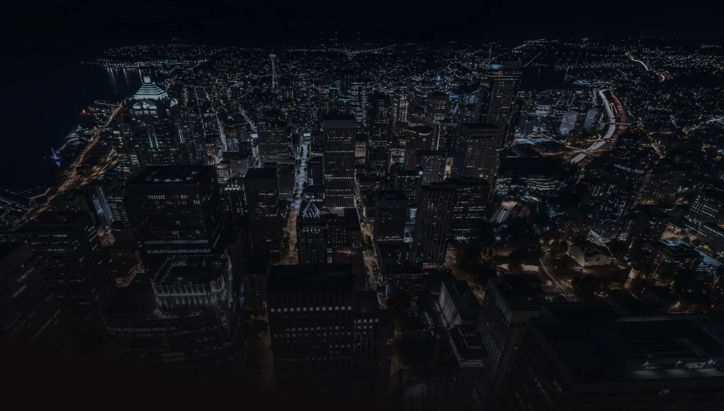 Aerial night view of a city with illuminated skyscrapers and streets, showing a busy urban landscape.