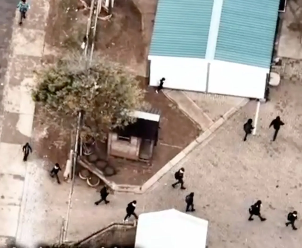 Aerial view of a group of people walking on a paved and dirt area near buildings and a tree.