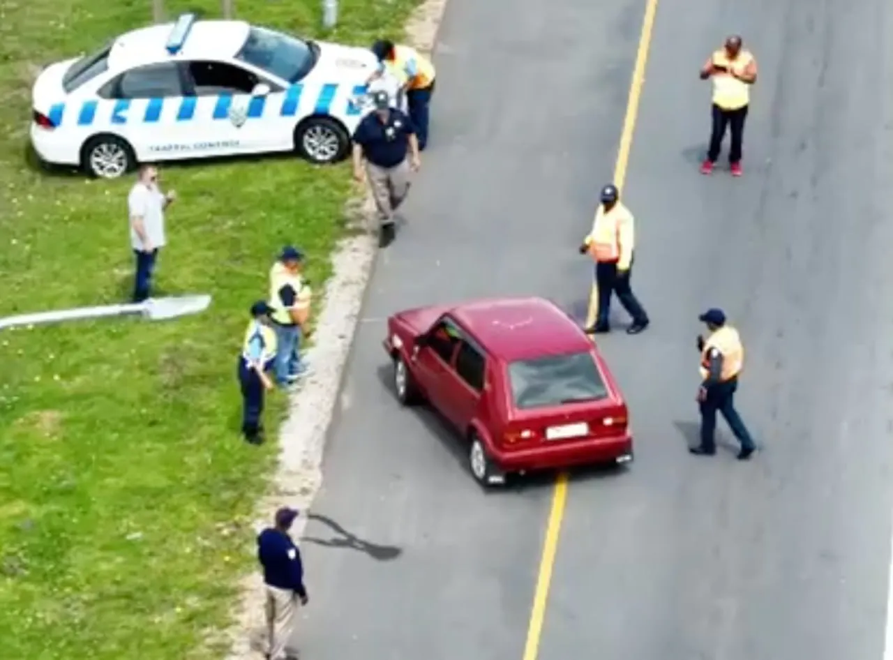 Red car stopped on road surrounded by traffic officers wearing yellow vests and a white traffic control car on the grass beside the road.