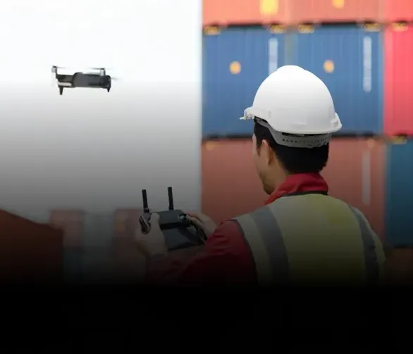 Worker in a hard hat and safety vest remotely controlling a flying drone near shipping containers.