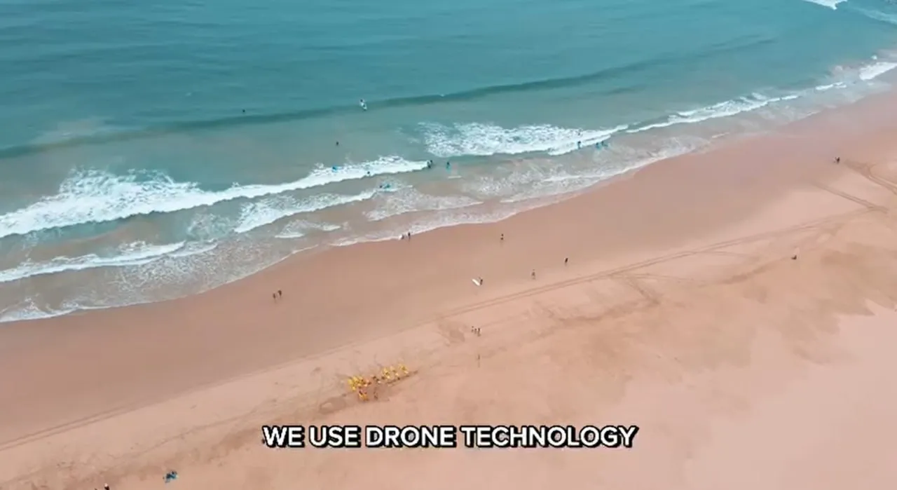 Aerial view of a sandy beach with scattered people near the shoreline and small waves in the ocean, overlaid with text 'WE USE DRONE TECHNOLOGY'.