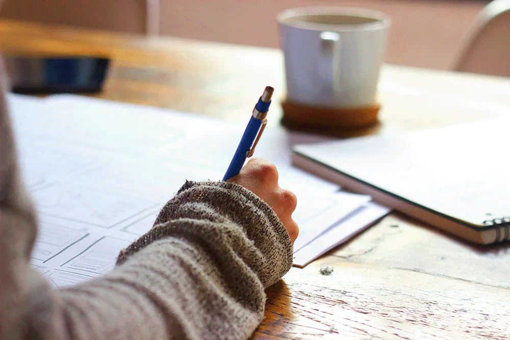Person writing with a blue pen on paper with documents and a cup of coffee on a wooden table.