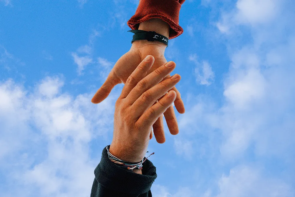 Two hands reaching and holding each other against a blue sky with scattered clouds.