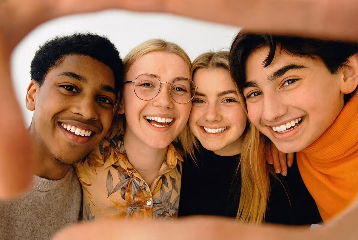 Four young adults smiling closely together while taking a selfie with hands framing the shot.