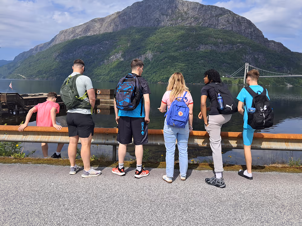 Group of six young people with backpacks standing and sitting by a guardrail overlooking a lake with mountains and a suspension bridge in the background.