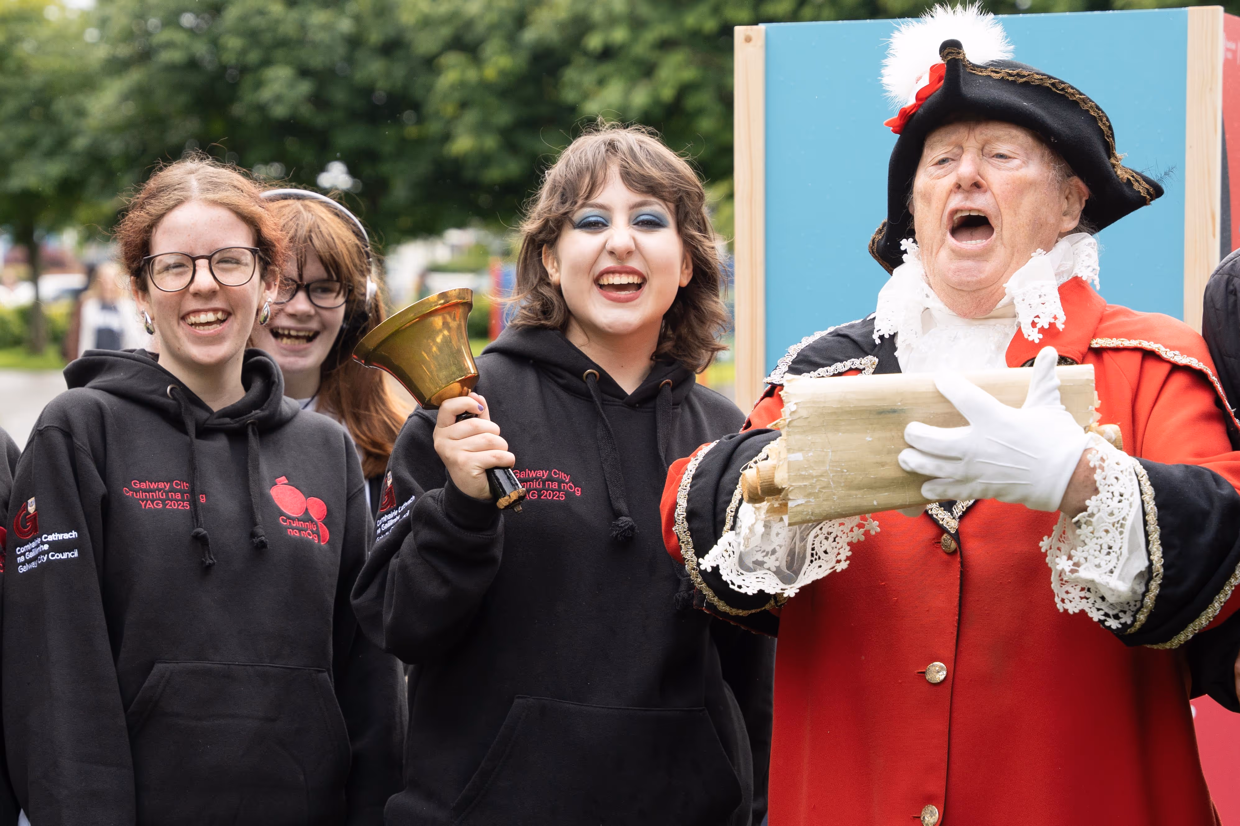 Three young women in black hoodies smiling, one holding a bell, next to a man in a red historical costume reading from a scroll.