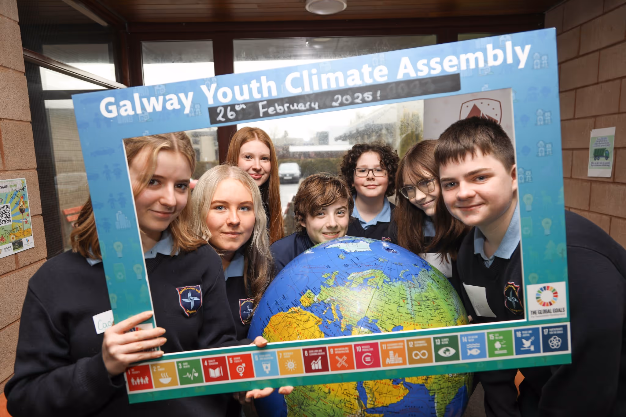 Seven youth in school uniforms holding a large globe and a frame reading 'Galway Youth Climate Assembly 26th February 2025' with UN Sustainable Development Goals icons along the bottom.