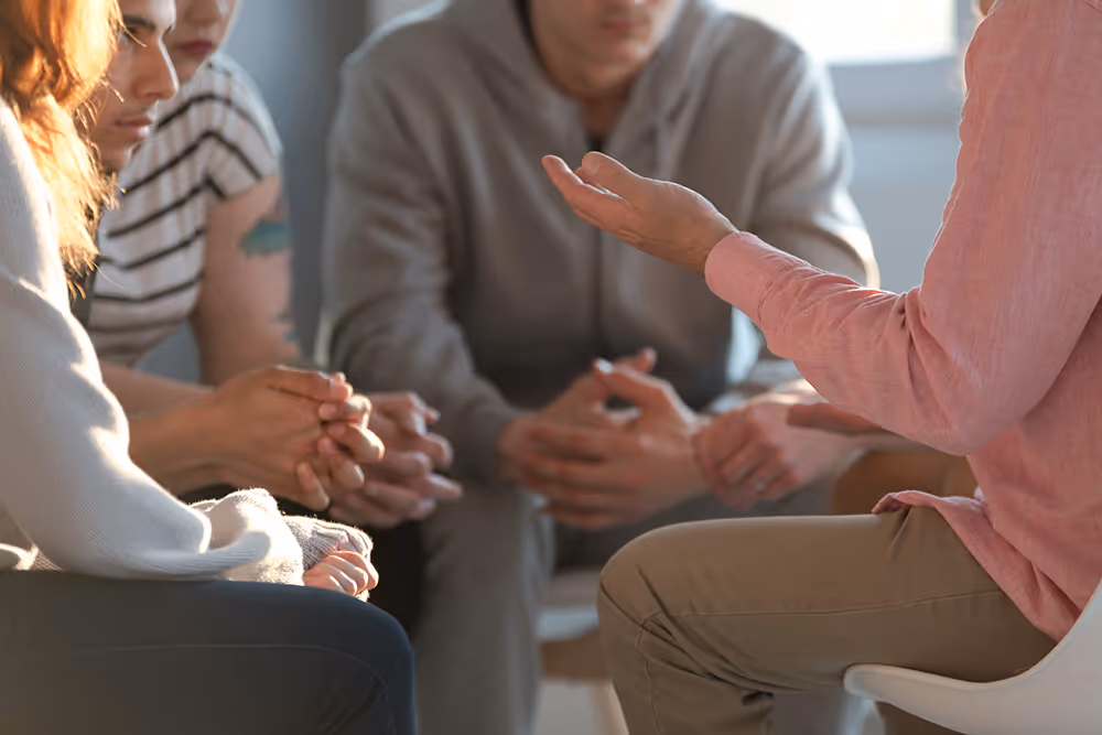 Group of young people sitting in a circle, listening attentively to a person speaking and gesturing with their hand.