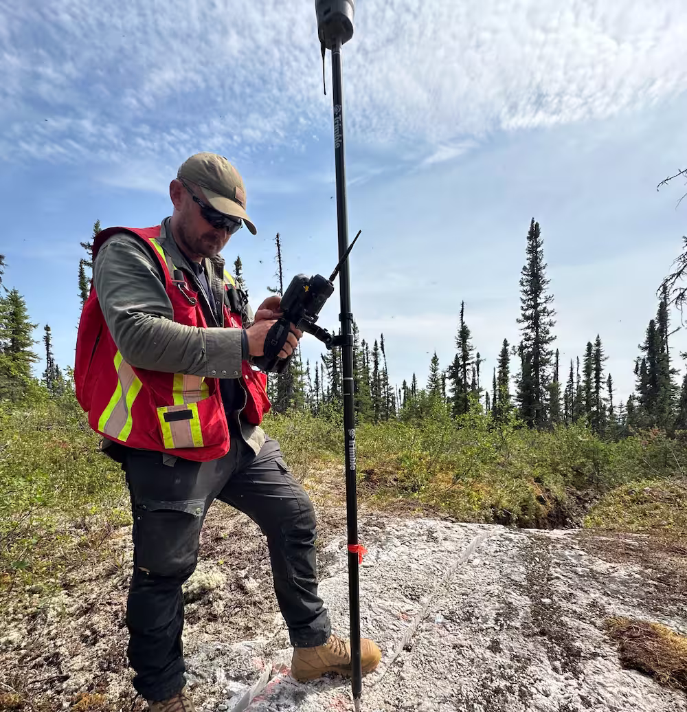 Man in safety vest using surveying equipment in a forested area under a partly cloudy sky.