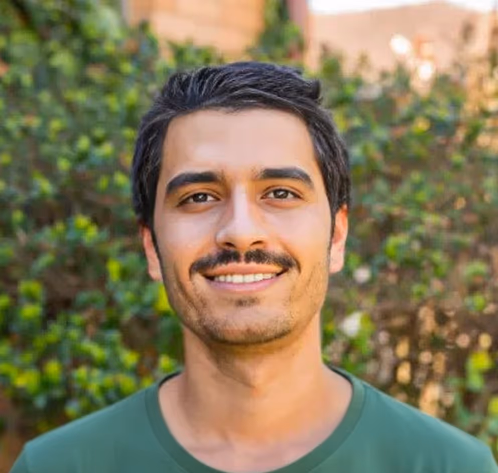 Smiling young man with dark hair and mustache wearing a green shirt, standing outdoors with greenery in the background.