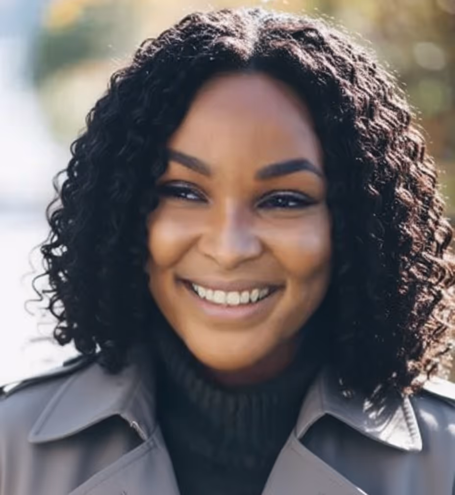 Smiling woman with curly hair wearing a gray coat and black turtleneck outdoors.