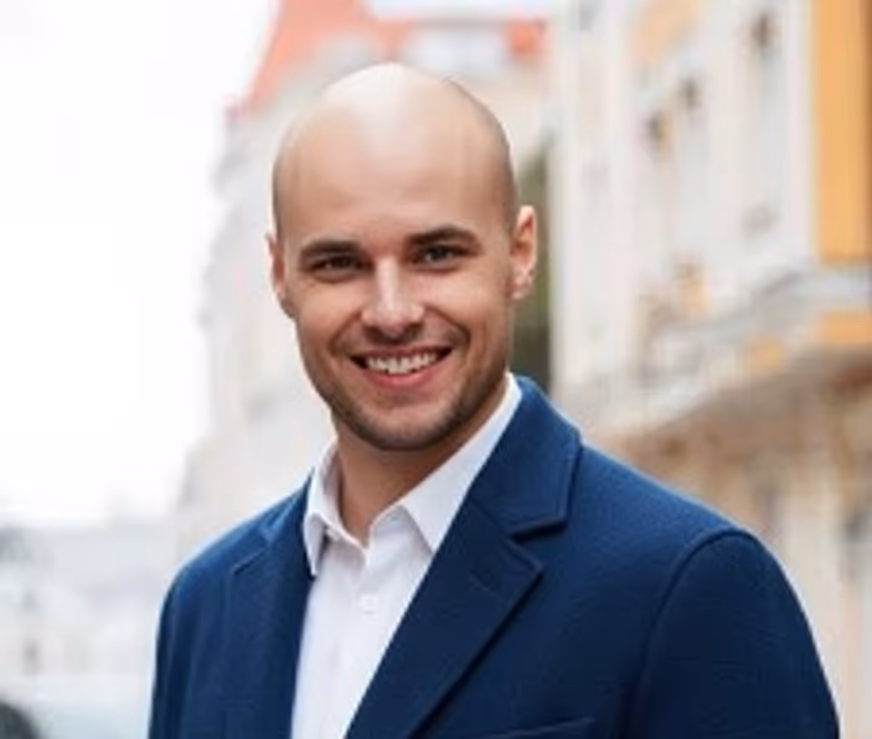 Smiling bald man wearing a white shirt and blue blazer outdoors with blurred buildings in the background.