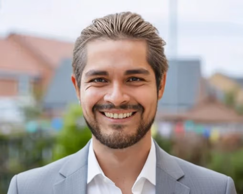 Smiling man with light brown hair and beard wearing a gray blazer and white shirt outdoors.