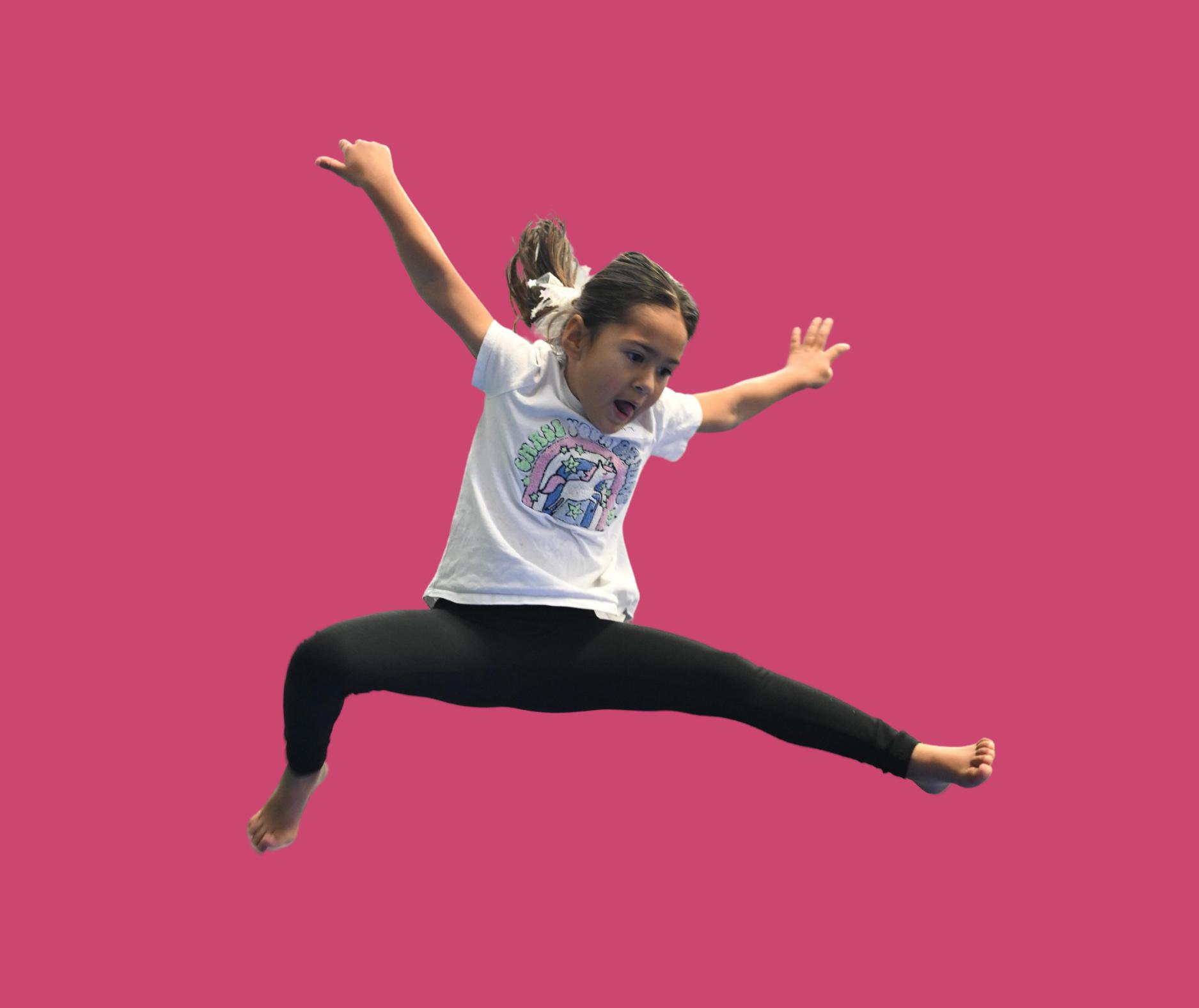Young girl in white t-shirt and black leggings jumps with legs wide apart against pink background.