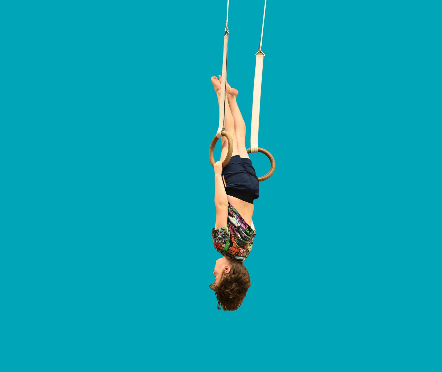 Child hanging upside down gripping wooden gymnastic rings against a plain turquoise background.