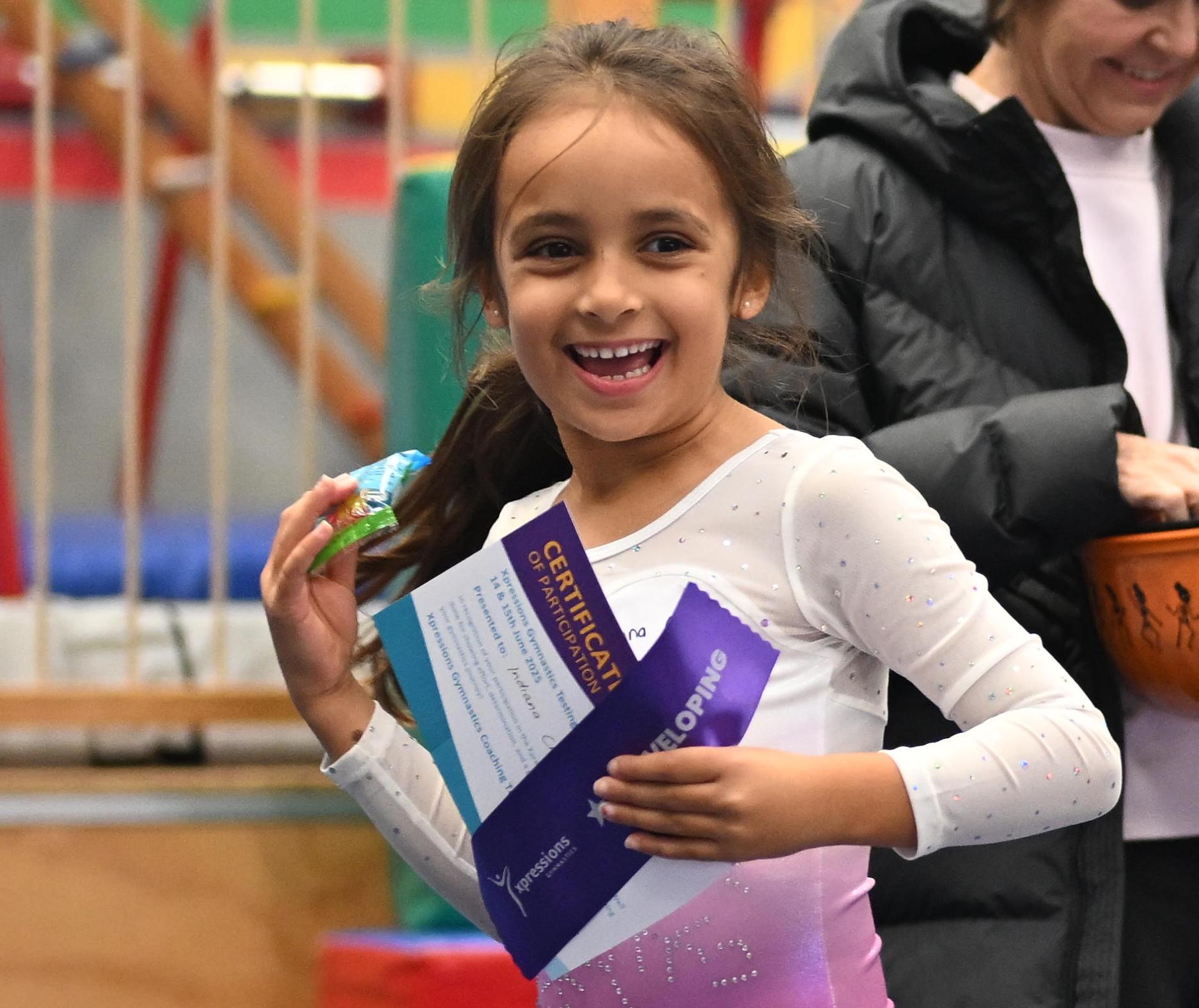 Smiling girl in gymnastics outfit holding certificate and ribbon at an indoor gym event.