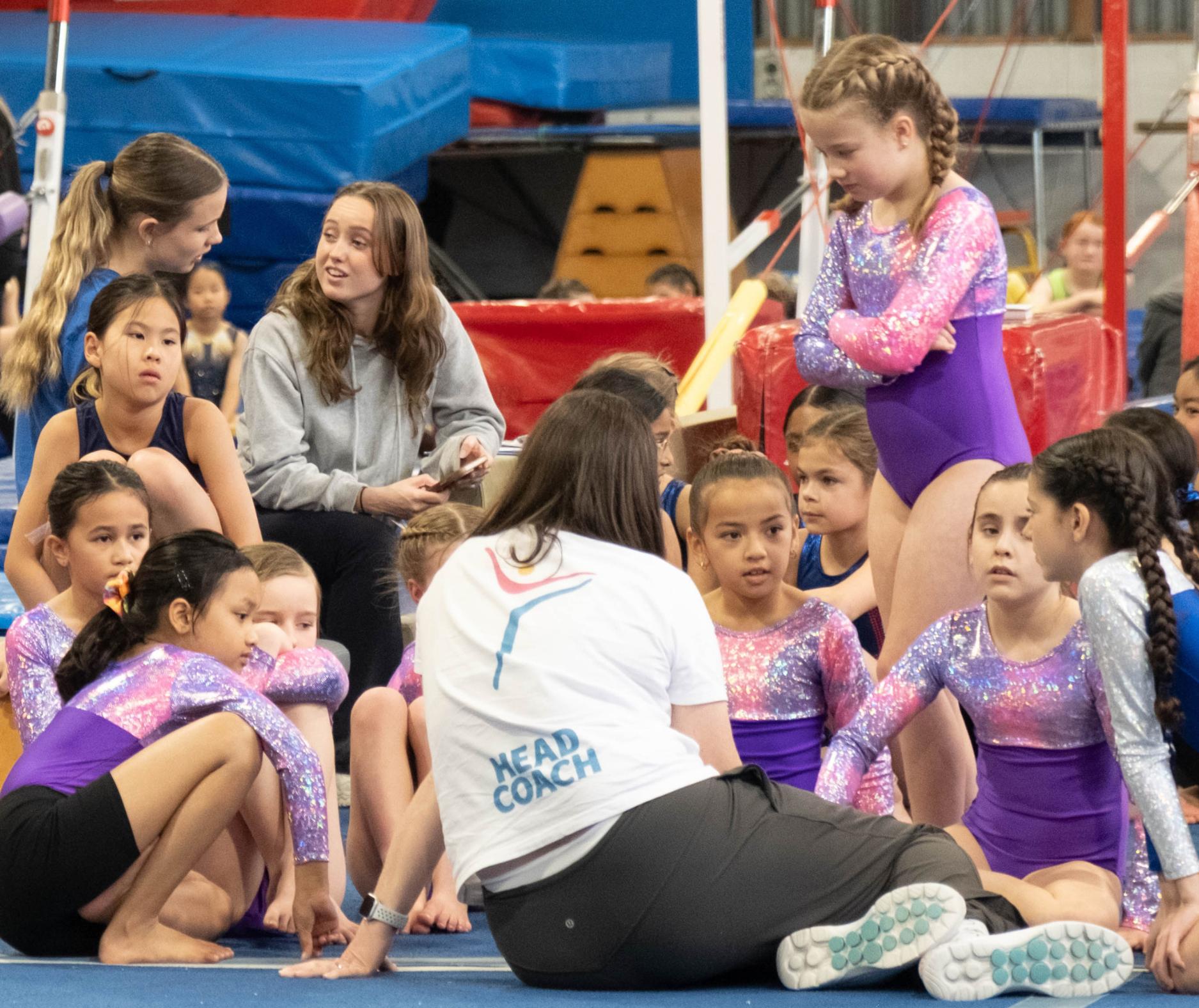 Young gymnasts in purple leotards listen to their head coach sitting on a gym floor during training.