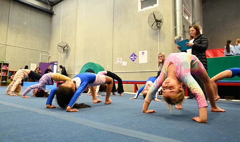Toddlers in colourful clothes doing backbends on blue mats during a gym class indoors.