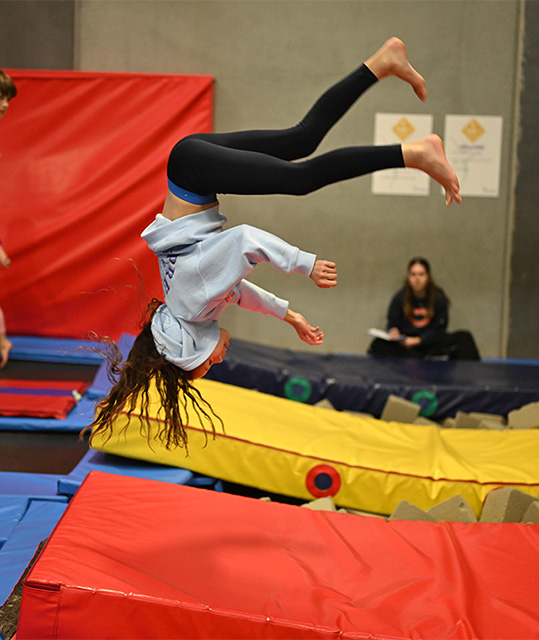 Girl with long hair in a hoodie mid-flip above padded mats in a gymnasium.
