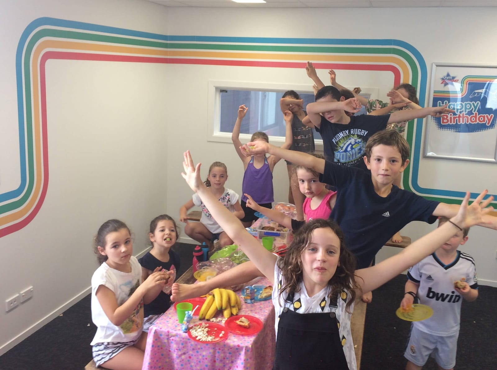 Group of children posing and celebrating with food and bananas on a table with a Happy Birthday sign.