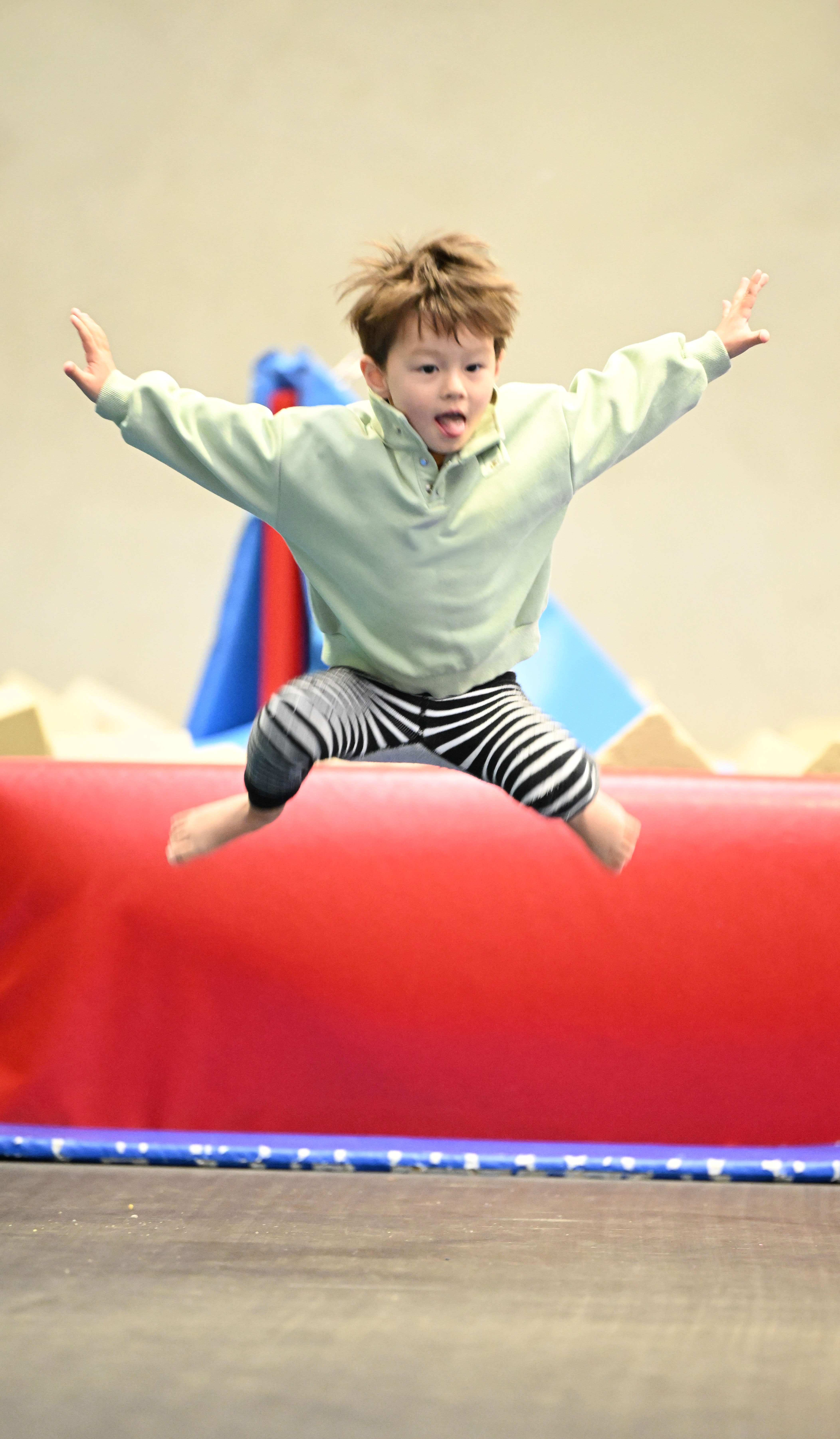 Young boy jumping with arms outstretched indoors on a trampoline with a red padded barrier.