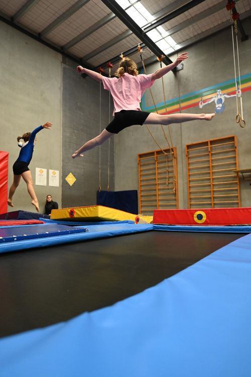 Two girls in a gym jumping on trampolines, one mid-air doing a split leap wearing pink and black.