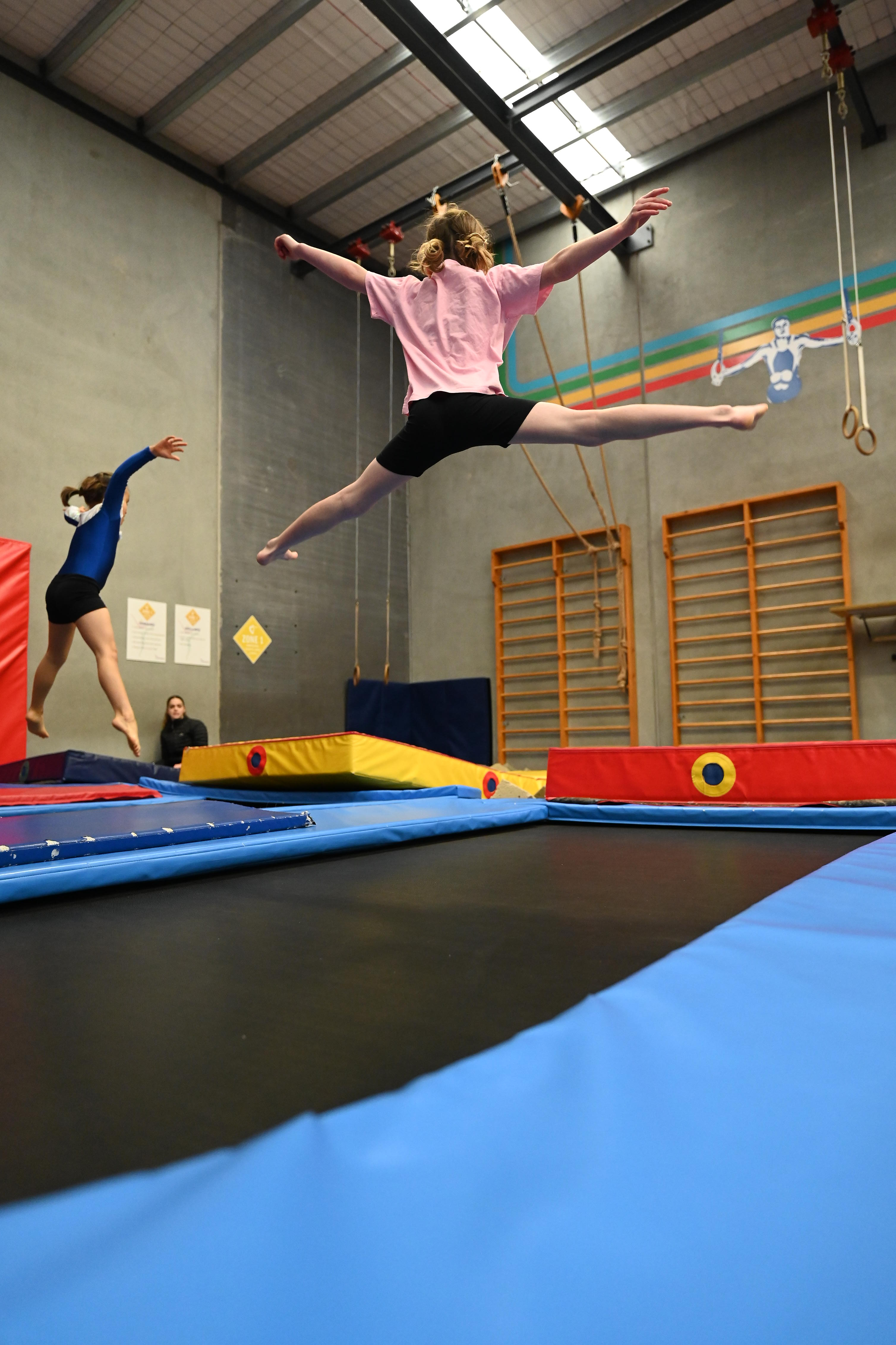 Two girls in a gym jumping on trampolines, one mid-air doing a split leap wearing pink and black.