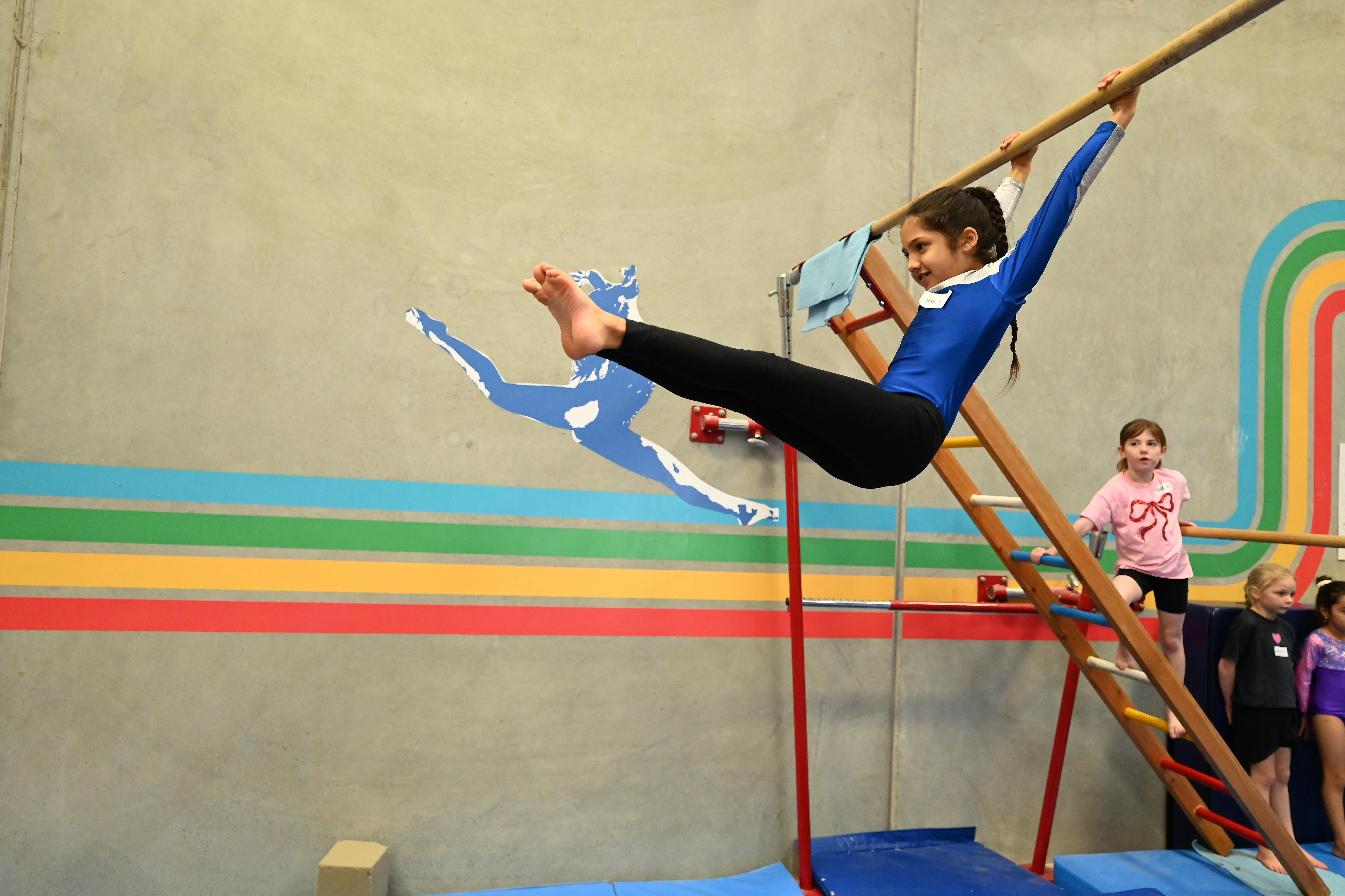 Girl in blue leotard holding gym bar with legs extended, other children wait in gymnasium.