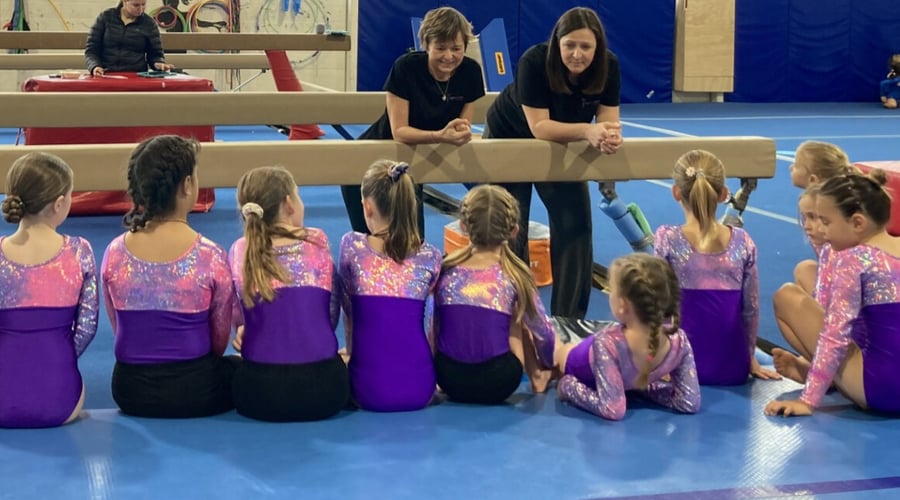 Young gymnasts in purple leotards sitting on floor listening to two female coaches at a balance beam.