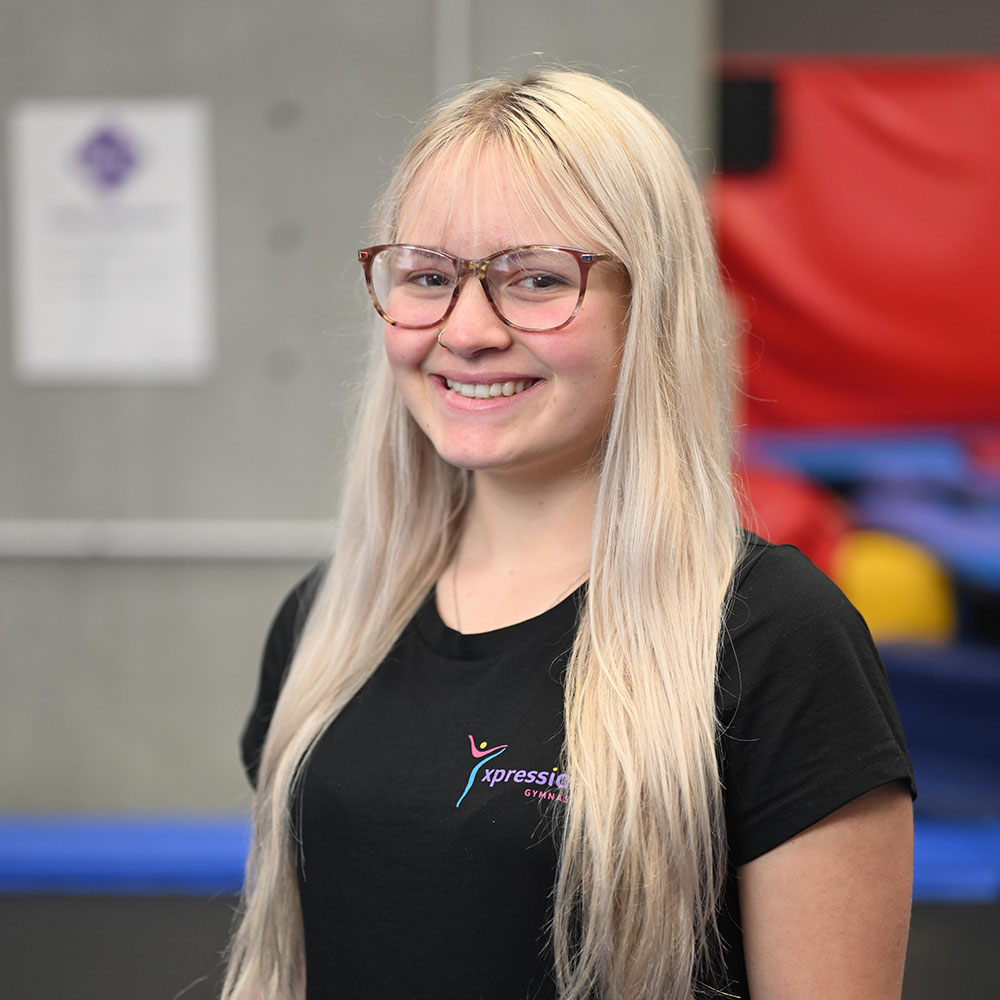 Smiling young woman with glasses and long blonde hair wearing a black Xpression Gymnastics t-shirt.