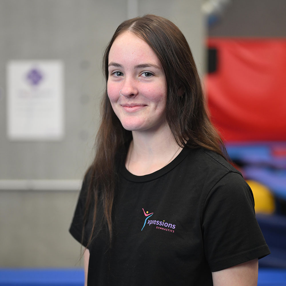Young woman with long brown hair and nose ring smiling, wearing black Xpressions Gymnastics t-shirt.