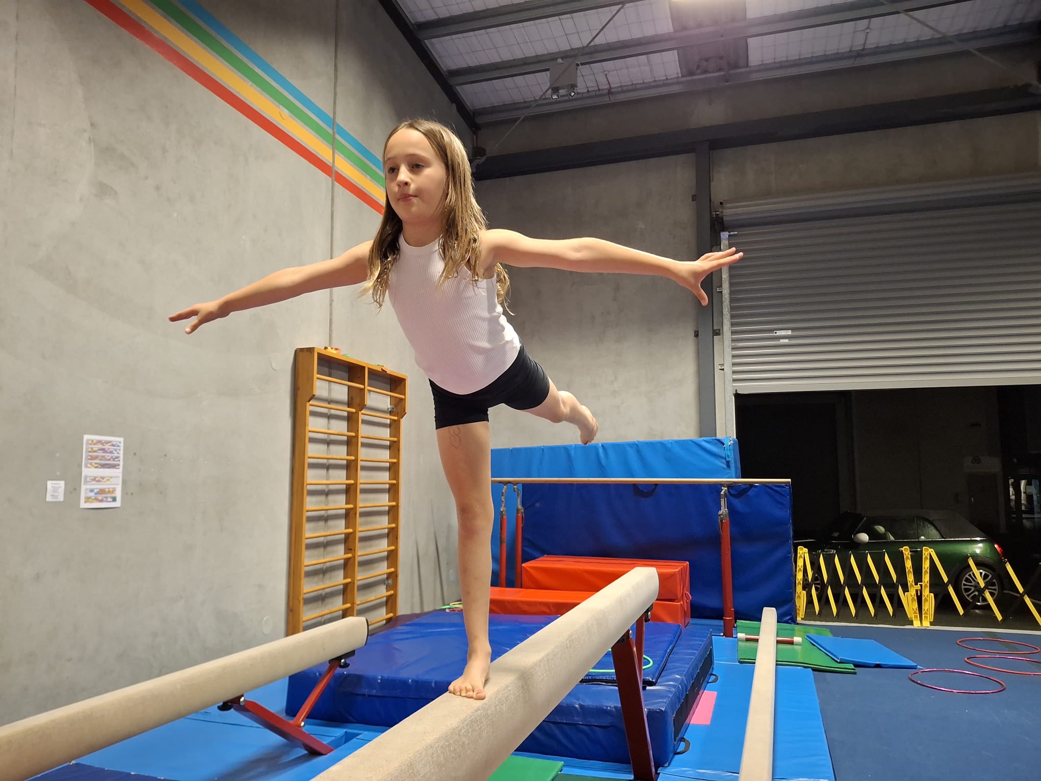 Young girl balancing on one foot on a gymnastics beam in a gym with mats and equipment.