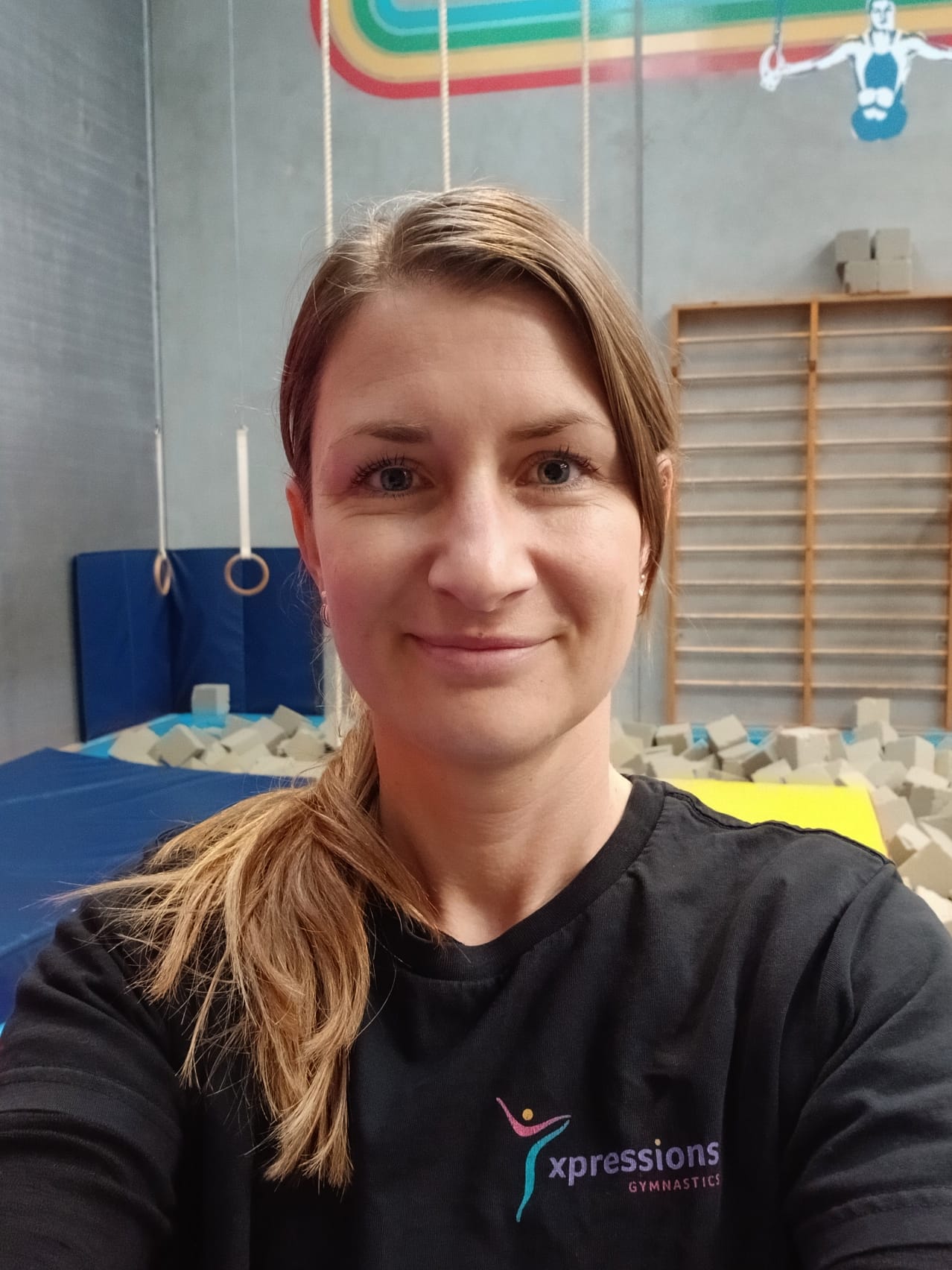 Woman with light brown hair in a ponytail wearing a black Xpressions Gymnastics shirt in a gym.