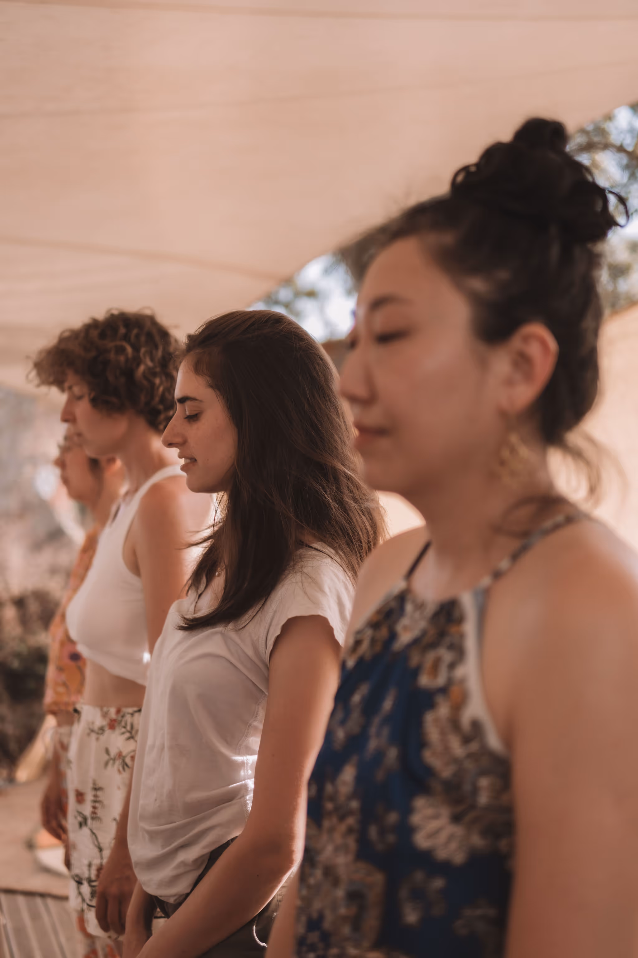 A group of women stand in a line under soft, natural light, all with eyes closed and relaxed expressions, suggesting they are meditating or participating in a mindful activity. They are dressed in casual summer clothing, with a neutral canopy overhead and trees faintly visible in the background.
