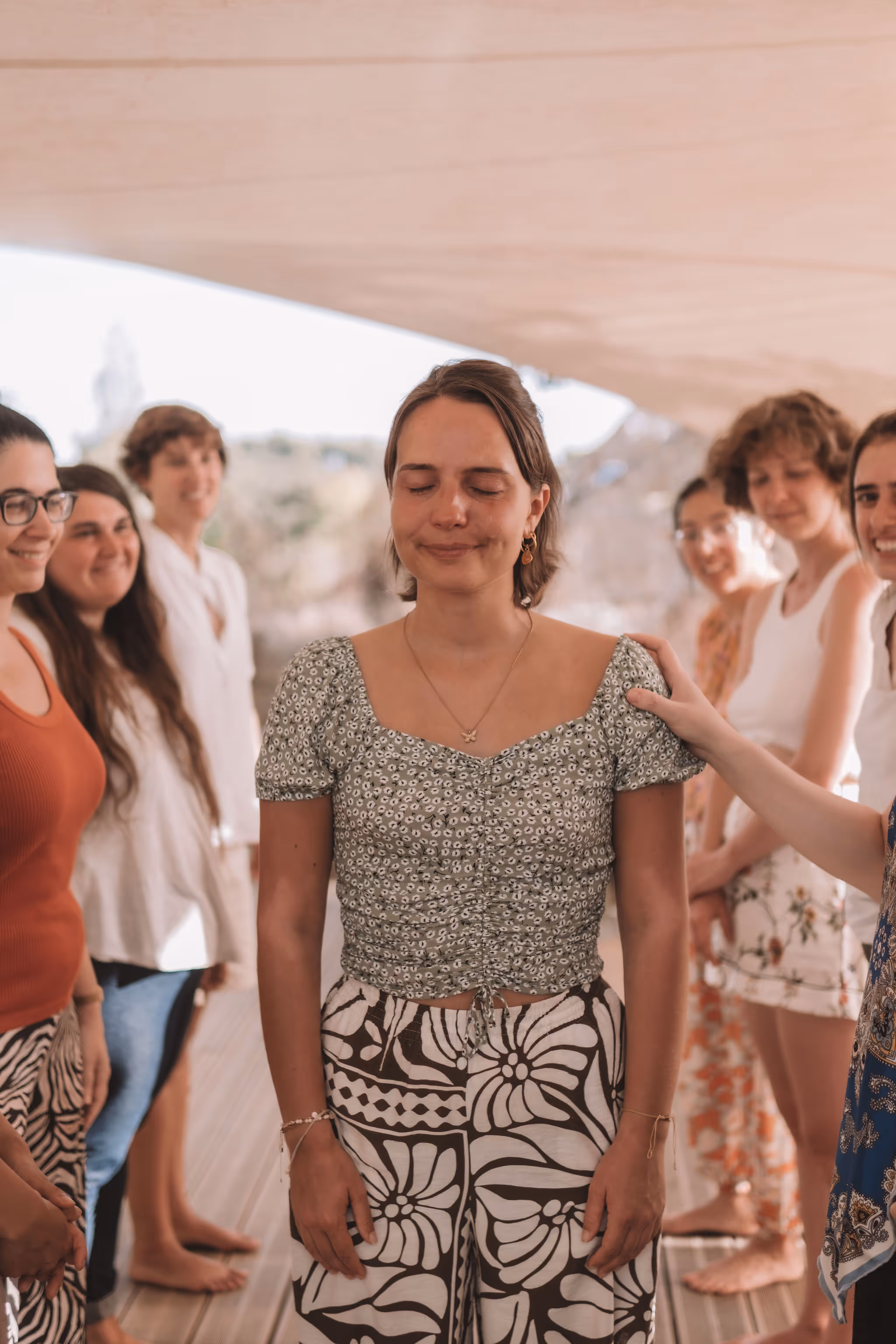 A group of women stands in a close circle under a canopy, gently placing their hands on one woman in the center who has her eyes closed and a peaceful smile. The moment appears heartfelt and supportive, suggesting a ritual of connection, affirmation, or emotional healing during a retreat.