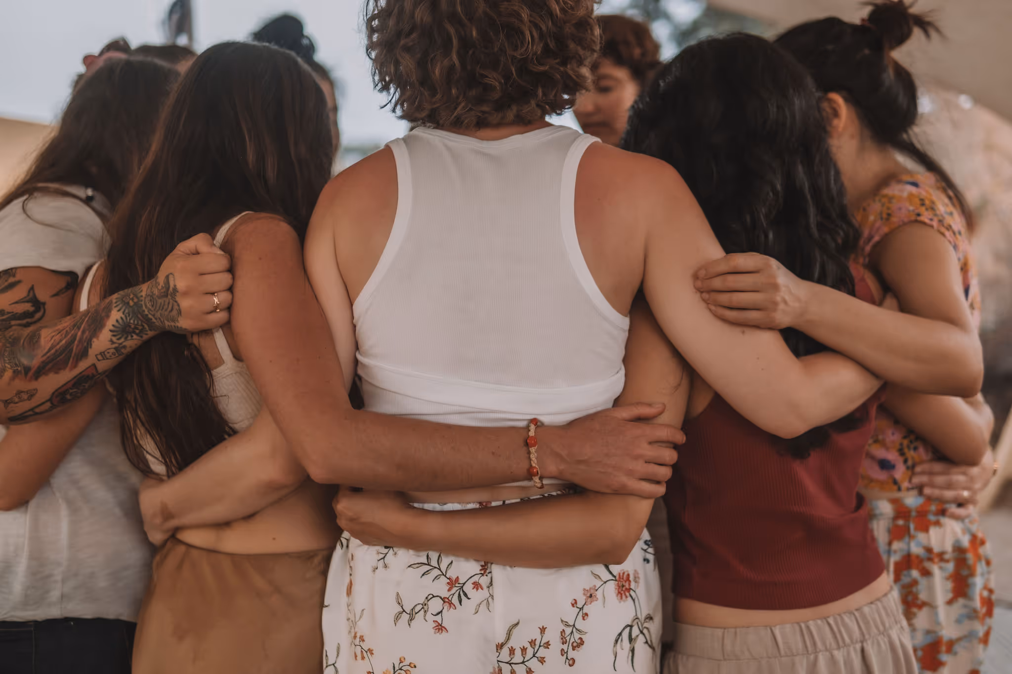 A close-up view of a group hug shows several people embracing tightly, their arms wrapped around one another in a supportive, unified circle. The photo captures a sense of intimacy and community, with floral clothing and visible tattoos adding personal detail.