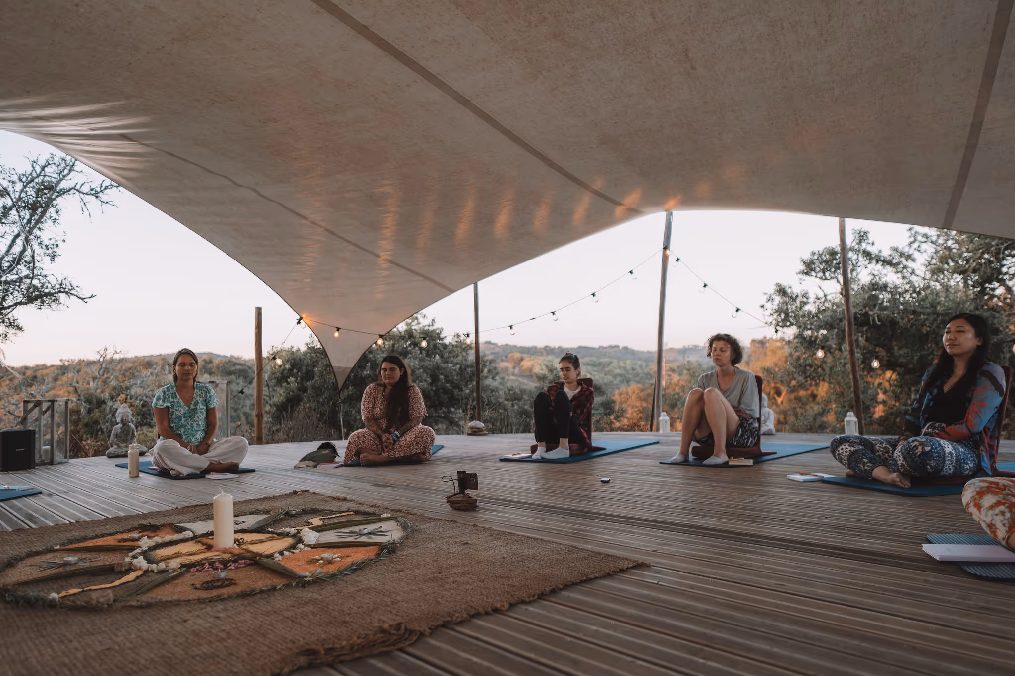 A group of women sits in a semi-circle on yoga mats under a large canopy, surrounded by nature and warm string lights. At the center is a decorative mandala made of natural materials with a white candle, suggesting a peaceful setting for meditation, reflection, or ceremony.